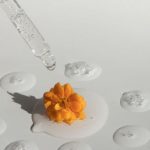 Essential Peripherals - From above of drops of transparent moisturizing cosmetic product dripped by pipette and small fresh flower head placed on white table