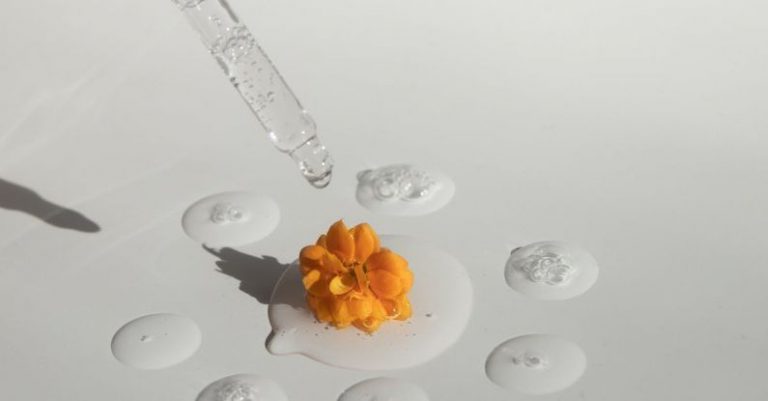 Essential Peripherals - From above of drops of transparent moisturizing cosmetic product dripped by pipette and small fresh flower head placed on white table